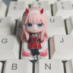 May include: A custom keycap featuring a pink-haired anime character with red horns and teal eyes. The keycap is in the shape of the character and is set on a white keyboard. The character is wearing a red outfit with black boots.