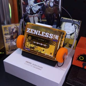 May include: A unique, gold-colored cassette tape-shaped headphone set with orange ear pads. The text "ZENLESS" is visible on the cassette. The headphones sit on a white box with the text "KMIDO Audio Studio CASSETTE @PLAYER".