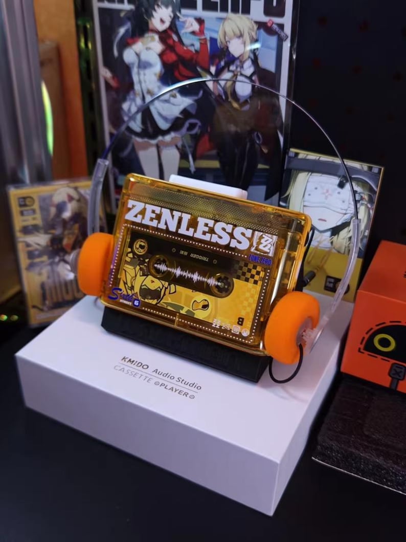 May include: A unique, gold-colored cassette tape-shaped headphone set with orange ear pads. The text "ZENLESS" is visible on the cassette. The headphones sit on a white box with the text "KMIDO Audio Studio CASSETTE @PLAYER".