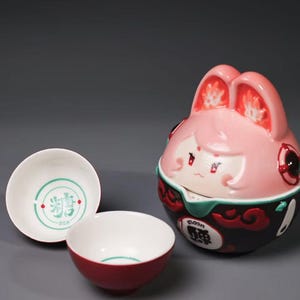 May include: A pink ceramic container with a cartoon face and rabbit ears, next to two white and red ceramic bowls. One bowl has green text and a design. The other bowl is red. The container has black and green accents.