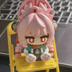 Könnte beinhalten: Eine rosa Anime-Figur mit großen Ohren und roten Augen, die auf einem kleinen gelben Stuhl sitzt. Die Figur trägt ein grün-schwarzes Outfit mit einem kreisförmigen Design. Der Stuhl hat ein schlichtes, minimalistisches Design.