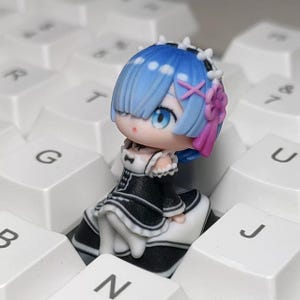 Może przedstawiać: Szczegółowa figurka postaci z anime z niebieskimi włosami, niebieskimi oczami i czarno-białą sukienką. Figurka ma różową kokardę i siedzi na białej klawiaturze. Klawisze klawiatury są widoczne w tle.