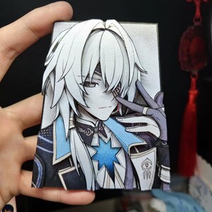 May include: A detailed paper cutout of an anime character with white hair and a blue and black outfit. The character has a star-shaped design on the chest and is making a peace sign with their fingers. The artwork is held in a hand.