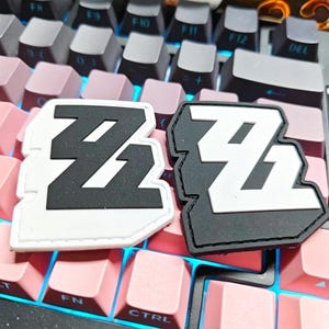 Op de afbeelding: Twee patches met het nummer "21" in een gestileerd lettertype. Eén patch is wit met zwarte details, de andere is zwart met witte details. De patches liggen op een toetsenbord met roze en grijze toetsen.