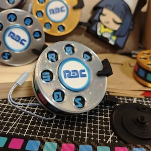 May include: A silver and blue retractable keychain with a circular design and the word "RAC" printed on it. The keychain has a black plastic clip and a silver string.