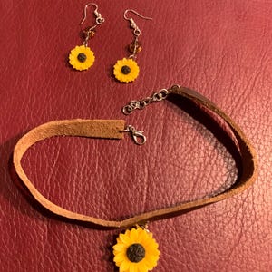 May include: A brown leather choker necklace with a sunflower charm. The necklace is paired with a set of sunflower earrings with amber glass beads.