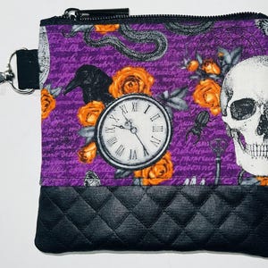 May include: A purple zippered pouch with a black quilted bottom. The front of the pouch features a skull, a clock, a raven, and orange roses on a purple background with a script pattern.