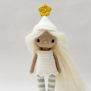 May include: A white crocheted doll with long blonde hair, wearing a white dress, grey and white striped leggings, and a white hat with a yellow star on top.