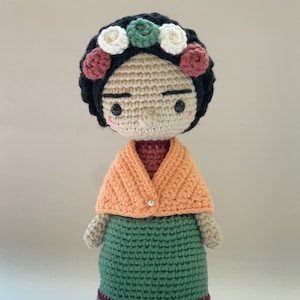 May include: Crocheted doll with black hair, a flower crown, an orange shawl, and a green dress. The doll is wearing a pearl button on the shawl. The doll is standing on a white surface. The text "Amour Fou" is visible in the bottom right corner of the image.