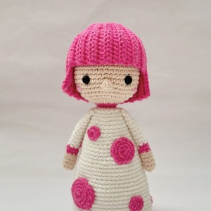 May include: A crocheted doll with pink hair and a white dress with pink polka dots. The doll is standing on a white background.