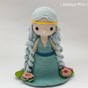 May include: A crocheted doll with long blue hair, wearing a teal dress and a yellow crown. The doll is standing on a bed of green lily pads with pink flowers.