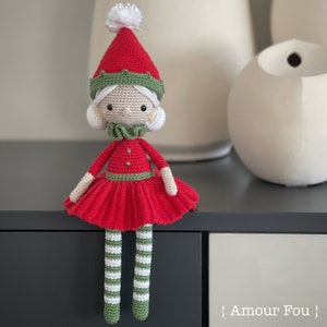 Op de afbeelding: Een gehaakte elfpop die een rode jurk, groen-wit gestreepte kousen en een groen-rode muts met een witte pompon draagt. De pop zit op een donkergrijs oppervlak.