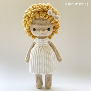 Op de afbeelding: Een gehaakte pop met blond krullend haar, die een witte jurk en een wit strikje draagt. De pop heeft een lieve uitdrukking en staat op een witte achtergrond.