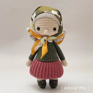 Op de afbeelding: Een gehaakte pop met een groen-wit gepatroneerd hoofddoek, een gele sjaal, een groene trui, een roze rok en bruine schoenen. De pop heeft een wit gezicht met zwarte ogen en een bruine neus.
