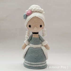 May include: Crocheted doll in a historical costume. The doll has a white bonnet with pink and blue floral accents, a gray dress with white trim, and curly white hair. The doll has a black choker and rosy cheeks. The doll is handmade.