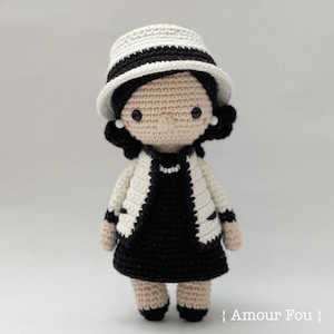May include: A crocheted doll wearing a white and black hat, a white and black jacket, and a black dress. The doll has black hair and a neutral facial expression. The doll is standing on a white background.