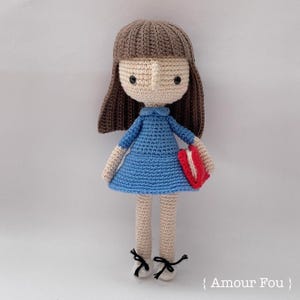 May include: A crocheted doll with brown hair, a blue dress, and a red book. The doll is wearing black shoes with white laces. The doll is standing on a white background. {Amour Fou}