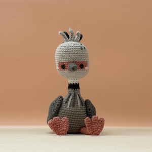 May include: A handmade crochet bird doll in shades of gray, black, and pink. The bird has a round head with a pink mask, black eyes, and a tuft of hair. The doll is sitting with its feet forward.