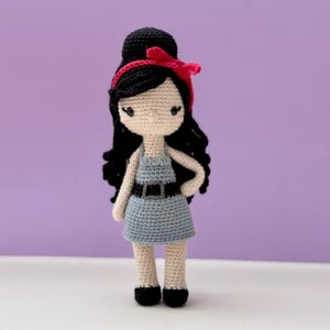 Może przedstawiać: Ręcznie robiona szydełkowa lalka z czarnymi włosami upiętymi w kok i loki, ubrana w jasnoniebieską sukienkę z czarnym paskiem. Lalka ma czerwoną opaskę na głowę i czarne buty. Lalka stoi na białej powierzchni na fioletowym tle.