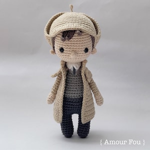 May include: A crocheted doll wearing a beige coat, a brown hat, and black pants. The doll has a beige face with black eyes and a brown hair.