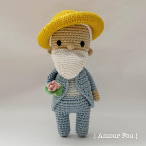 Puede incluir: Un juguete de peluche tejido a crochet que representa a una persona con un sombrero amarillo, barba blanca, traje azul y sosteniendo una flor rosa. El juguete está de pie sobre un fondo blanco. El texto "Amour Fou" es visible en la esquina inferior derecha de la imagen.