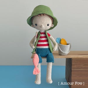 Peut inclure: Poupée au crochet faite à la main représentant un garçon pêcheur. La poupée porte un chapeau vert, un gilet et un short bleu. Il tient une canne à pêche avec un poisson rose. Un petit seau avec de la laine colorée est à côté de lui.