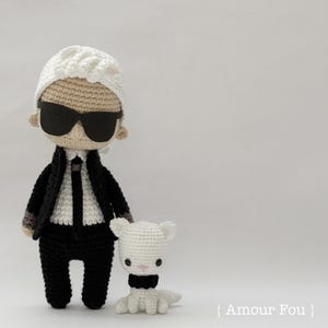 May include: A crocheted doll wearing a black suit, white shirt, and sunglasses. The doll is standing next to a smaller crocheted white cat wearing a black bow tie. The text "{ Amour Fou }" is visible in the bottom right corner of the image.