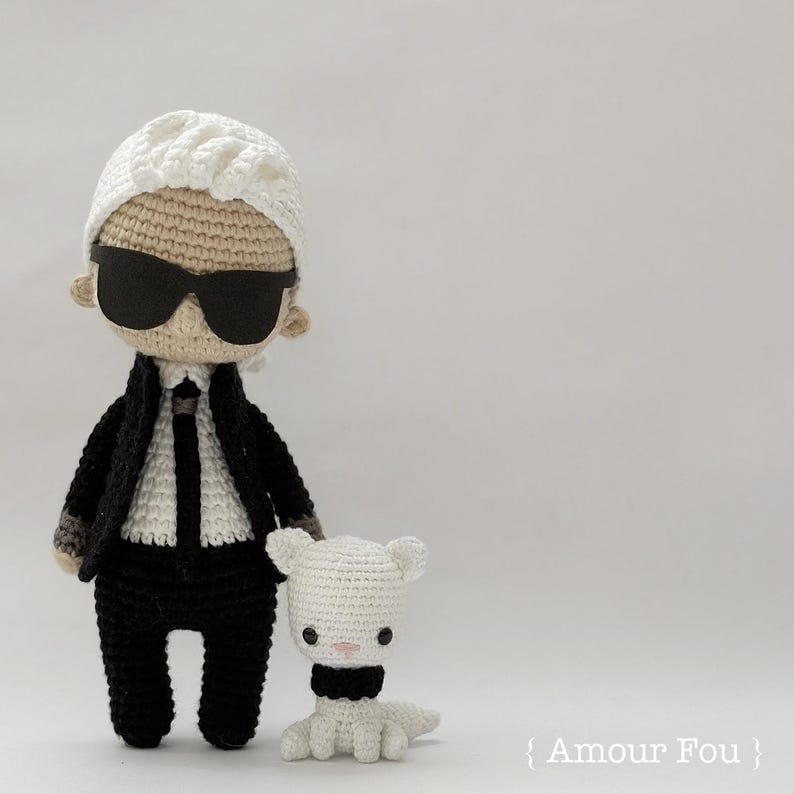 May include: A crocheted doll wearing a black suit, white shirt, and sunglasses. The doll is standing next to a smaller crocheted white cat wearing a black bow tie. The text "{ Amour Fou }" is visible in the bottom right corner of the image.