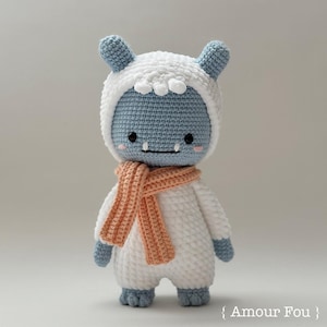 May include: A crocheted plush toy with a white body, blue head, and orange scarf. The toy has a friendly face with black eyes and a toothy grin.