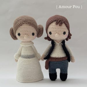 May include: Two crocheted dolls, one with a white dress and brown hair, the other with brown hair and a brown vest, blue pants, and a brown belt. The dolls are standing on a white surface. The text "Amour Fou" is written in black on a white background above the dolls.