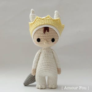 Puede incluir: Una muñeca amigurumi de crochet blanco que lleva una corona de crochet amarilla con cuernos. La muñeca tiene el pelo de lana marrón y ojos de botón negros.