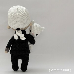 May include: A crocheted doll wearing a black suit and a white hat, holding a white crocheted animal. The doll is standing on a white background.