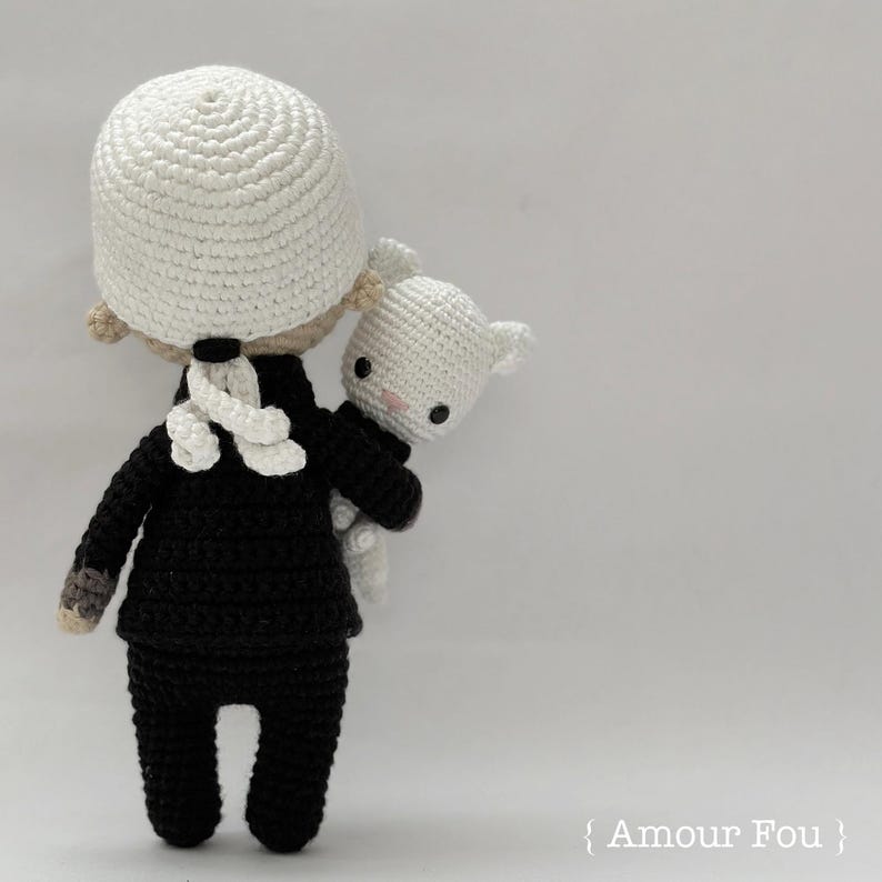May include: A crocheted doll wearing a black suit and a white hat, holding a white crocheted animal. The doll is standing on a white background.