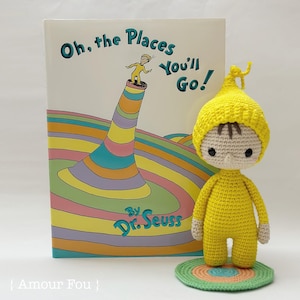 May include: A yellow crocheted amigurumi doll wearing a yellow hat, standing on a colorful crocheted mat. The doll is based on the character from the book "Oh, the Places You'll Go!" by Dr. Seuss.
