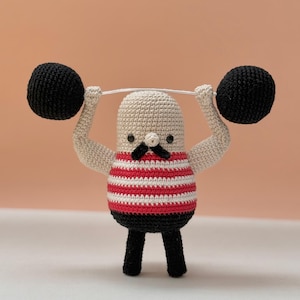 May include: A crocheted figurine of a weightlifter with a beige head and torso, a black mustache, and black eyes. The figure wears a red and white striped shirt and black pants, while lifting a barbell with two large black crocheted weights.