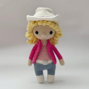 May include: A crocheted doll featuring blonde curly hair, a white cowboy hat, a bright pink jacket, a light pink shirt, and blue pants. The doll has black bead eyes and a light beige complexion, crafted with visible yarn stitches throughout the figure.