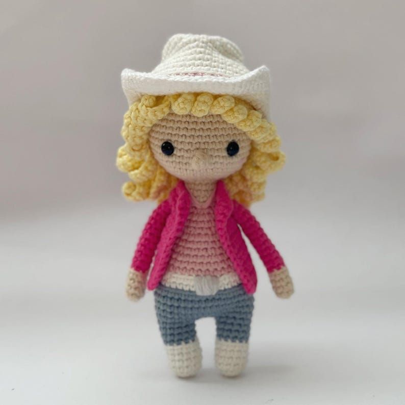 Peut inclure: Une poup&eacute;e crochet&eacute;e avec des cheveux blonds boucl&eacute;s, un chapeau de cowboy blanc, une veste rose vif, un haut rose clair et un pantalon bleu. La poup&eacute;e a des yeux en perles noires et un teint beige clair, r&eacute;alis&eacute;e avec des mailles visibles.