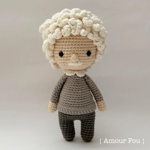 May include: A handmade crochet doll with a light skin tone, black button eyes, and a small white mustache. The doll has curly white hair, a gray sweater, and dark gray pants. The doll is standing on a white surface. The text "{ Amour Fou }" is in the bottom right corner.