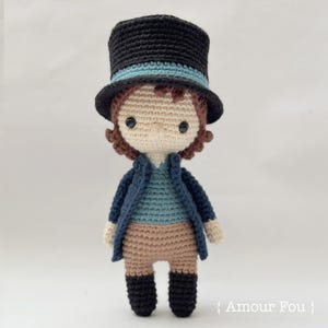 May include: A crocheted doll wearing a black top hat with a blue band, a blue shirt, a brown jacket, brown pants, and black boots. The doll has brown hair and a white face with black eyes.