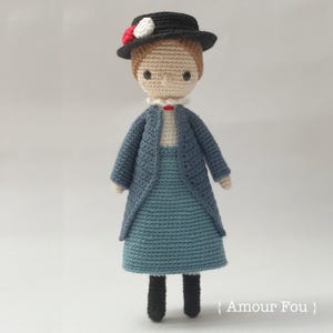 Puede incluir: Una muñeca de crochet que lleva un vestido azul, una chaqueta azul, un sombrero negro con flores blancas y rojas y zapatos negros. La muñeca está de pie sobre un fondo blanco. El texto "Amour Fou" es visible en la esquina inferior derecha de la imagen.
