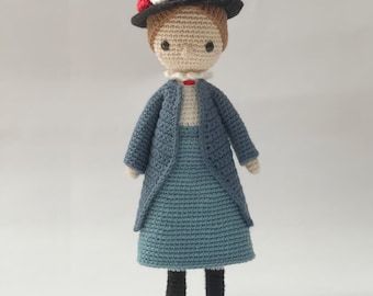 May include: A crocheted doll wearing a blue dress, a blue cardigan, a black hat with white and red flowers, and black shoes. The doll is standing on a white background. The text "Amour Fou" is visible in the bottom right corner of the image.
