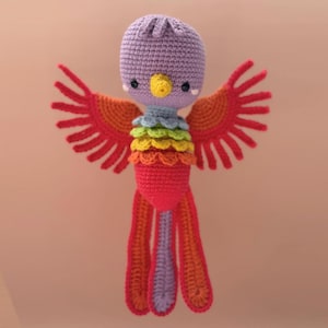 Könnte beinhalten: Ein handgefertigtes Häkeltier in Form eines Vogels mit lila Kopf, gelbem Schnabel und regenbogenfarbener Brust. Die Flügel sind rot und orange, die Schwanzfedern orange, lila und rot. Das Spielzeug hat ein verspieltes Design.
