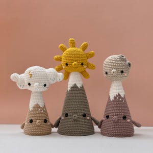 Op de afbeelding: Drie handgemaakte gehaakte speelgoedjes met een weer-thema. Het speelgoed omvat een zon, een wolk en een maan, elk bovenop een bergvormige basis. De zon is geel, de wolk is wit met een gouden bliksem en de maan is grijs.