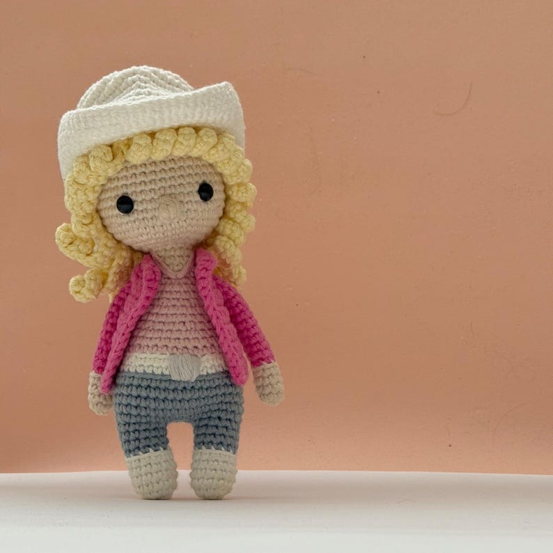 Peut inclure: Une poup&eacute;e crochet&eacute;e &agrave; la main avec un chapeau de cow-boy cr&egrave;me, des cheveux en laine blonde boucl&eacute;e et des yeux en perles noires. La poup&eacute;e porte une chemise rose clair, une veste ouverte rose vif, une ceinture blanche, un pantalon gris et des bottes cr&egrave;me.