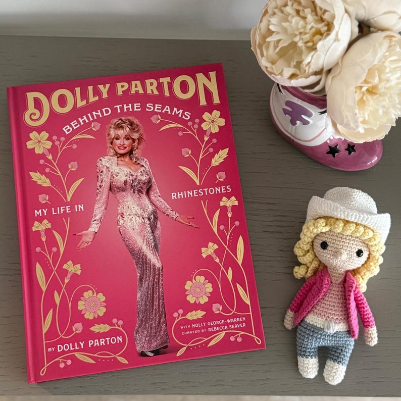 Peut inclure: Un livre reli&eacute; intitul&eacute; Dolly Parton Behind the Seams My Life in Rhinestones avec une couverture rose pr&eacute;sentant une photographie de Dolly Parton en robe &agrave; paillettes, accompagn&eacute; d'une petite poup&eacute;e crochet&eacute;e portant un chapeau de cow-boy blanc.