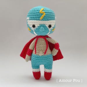 May include: A crocheted toy wrestler wearing a blue mask with a yellow lightning bolt, a red cape, and white and red boots.