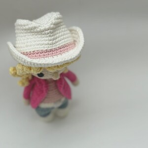 Peut inclure: Une petite poup&eacute;e au crochet avec des cheveux en laine blonde, un chapeau de cow-boy blanc avec un ruban rose clair, une veste rose vif, un haut ray&eacute; et un pantalon bleu. La figurine poss&egrave;de des yeux en perles noires et des mailles visibles.