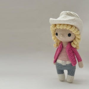 Peut inclure: Une poup&eacute;e en crochet portant un chapeau de cowboy cr&egrave;me, des cheveux jaunes boucl&eacute;s et des yeux en perles noires. La poup&eacute;e porte une veste rose vif sur un haut rose clair, une ceinture blanche, un pantalon gris et des bottes blanches.