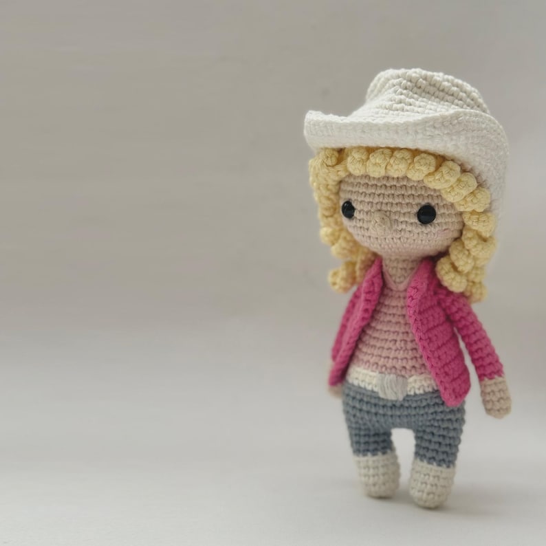 Peut inclure: Une poup&eacute;e en crochet portant un chapeau de cowboy cr&egrave;me, des cheveux jaunes boucl&eacute;s et des yeux en perles noires. La poup&eacute;e porte une veste rose vif sur un haut rose clair, une ceinture blanche, un pantalon gris et des bottes blanches.