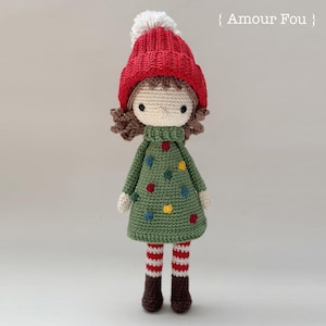 May include: A crocheted doll wearing a red hat with a white pom-pom, a green dress with colorful dots, and red and white striped leggings. The doll has brown hair and brown eyes.